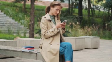 Güzel, şık, sarışın bir kız, akıllı telefonuyla arkadaşıyla sohbet ediyor, bankta oturuyor, parkta kitaplarıyla oturuyor. Açık hava konseptini çalış