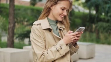 Parktaki bankta oturan, akıllı telefondan erkek arkadaşıyla sohbet eden ve gülümseyen neşeli öğrenci kızı yan bakışı.