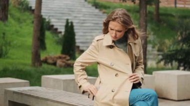 Trençkot giymiş genç bir kız öğrenci şehir parkındaki bankta akıllı telefondan konuşuyor.