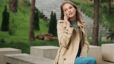 Trençkotlu güzel bir kızın, parkta bir bankta otururken, telefonda arkadaşlarıyla konuşurken görüntüsü.