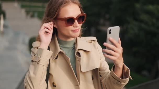 在户外用智能手机与男朋友视频交谈时 穿着太阳镜和风衣打扮的迷人的随和女孩 — 图库视频影像