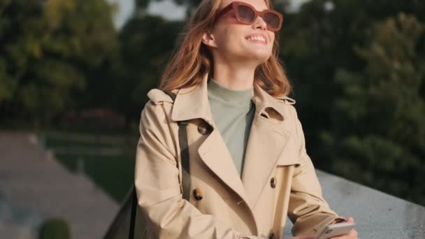 Hezké Usmívající Dívka Nosí Sluneční Brýle Trenč Kabát Drží Smartphone — Stock video