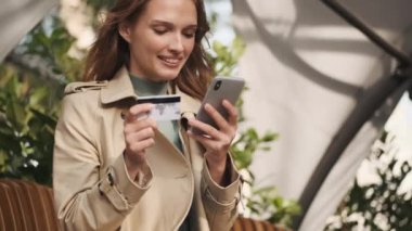 Çevrimiçi alışverişleri kredi kartıyla ödeyen çekici gülümseyen kız sokak kafesinde kahve molasında akıllı telefon kullanıyor.