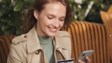 Güzel bir kız öğrencinin kredi kartıyla ödeme yapmak için akıllı telefonu kullanmasını ve açık kafede dinlenirken gülümsemesini görüntülemek. Modern teknoloji