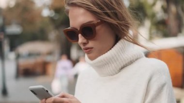 Güneş gözlüklü, beyaz, rahat bir kazak giymiş güzel bir kız. Erkek arkadaşıyla mesajlaşıp onu şehir caddesinde bekliyor.