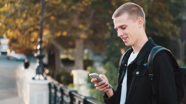 路上でスマートフォンを使って友達と幸せなテキストメッセージを探しているハンサムな学生の男 勉強の後で休んでいる若い学生 — ストック動画
