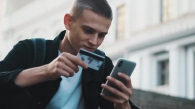 Çevrimiçi alışverişleri akıllı telefon ve kredi kartıyla ödeyen yakışıklı öğrenci portresi.