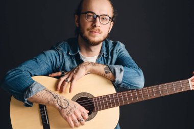 Young attractive tattooed man looking confident posing with guitar isolated on black background. Stylish musician with acoustic guitar