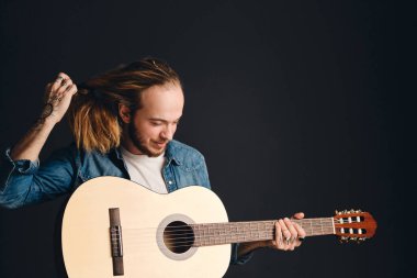 Young tattooed guy with guitar untying his hair isolated on black background. Male musician with acoustic guitar in studio