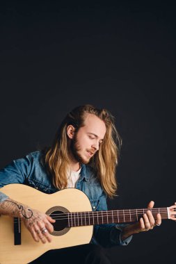 Side view of long haired tattooed male musician looking sensual playing on acoustic guitar over black background. Stylish singer with guitar