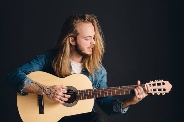 Side view of attractive long haired tattooed guy playing on acoustic guitar and singing preparing for concert isolated on black background. Stylish singer with guitar