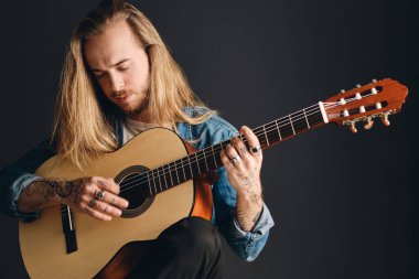 Attractive long haired tattooed man playing on acoustic guitar composing song isolated on black background. Stylish male singer with guitar