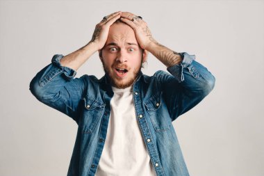 Portrait of young bearded man looking upset being amazed with negative news holding head with both hands over white background. Face expression