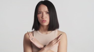 Ciddi bir Asyalı kız kızgın görünüyor. Hiçbir hareket göstermiyor. Ellerini çapraz tutarak beyaz arka plan üzerinde geziyor.