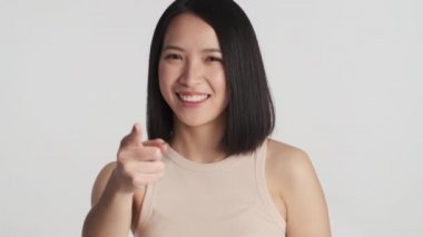 Çekici Asyalı kız, beyaz arka planda mutlu görünen işaret parmağıyla kameraya doğru bakıyor.