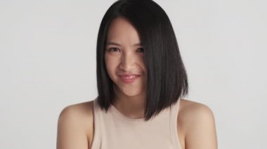Güzel Asyalı kız beyaz arka planda kamerada utangaç şirin görünüyor. Güzel kadın gülümseyerek yabancılardan iltifat alıyor.