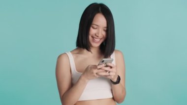 Güzel gülümseyen Asyalı kız mavi arka planda izole edilmiş akıllı telefondan sevgilisiyle mesajlaşırken mutlu görünüyor. Modern iletişim