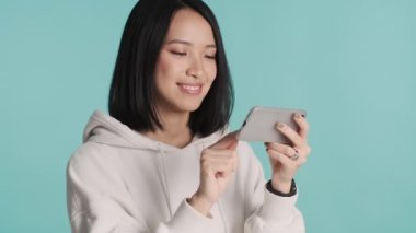 Kapüşonlu güzel, gülümseyen Asyalı kadın internette renkli arka plan üzerinden akıllı telefon kullanarak komik videolar izliyor.