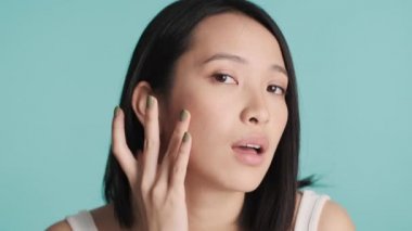 Mavi arka planda izole edilmiş makyaja hazırlanan krem sürdükten sonra genç ve çekici Asyalı kadın yüzüne dokunuyor. Güzellik konsepti