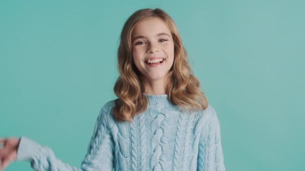 きれいなブロンドの波状の髪の十代の少女は 居心地の良いセーターの挨拶の友人に身を包んだとカラフルな背景にカメラに笑みを浮かべて チャーミング女の子手を振るこんにちは — ストック動画
