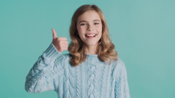 かなり肯定的なブロンドの十代の女の子は青の背景の上にカメラに親指を維持 表現のように — ストック動画