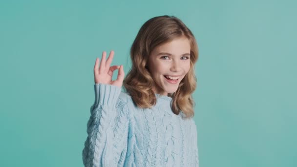 魅力的な肯定的なブロンドの十代の女の子は大丈夫と青の背景に隔離されたカメラに親指を維持します 表現のように ボディランゲージの概念 — ストック動画