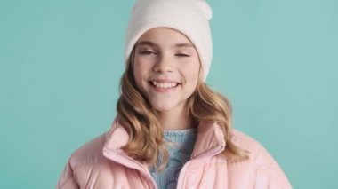 Pembe ceketli ve şapkalı hoş bir genç kız mavi arka planda mutlu görünüyor.