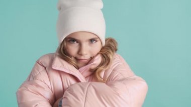 Pembe ceketli, şapkalı, üşümüş ama mavi arka planda gülümseyen güzel bir genç kız.
