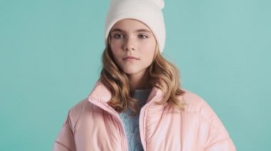 Çekici sarışın kız pembe ceketli ve şapkalı. Mavi arka planda izole edilmiş kameraya gülümsüyor.