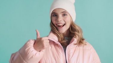 Pembe ceketli ve şapkalı güzel sarışın genç kız mavi arka planda parmağını yukarıda tutarak mutlu görünüyor. İfade gibi.