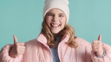 Pembe ceketli ve şapkalı neşeli sarışın kız mavi arka planda kameranın önünde başparmağını tutuyor. İfade gibi.