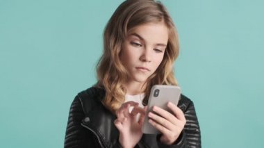 Ciddi sarışın genç kız, arkadaşından mesaj alıyor mavi arka planda akıllı telefondan sohbet ediyor. Tatminsiz yüz ifadesi