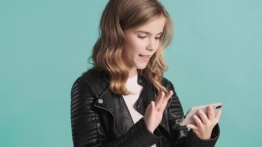 Deri ceketli sarışın genç kız akıllı telefondan videolar izliyor mavi arka planda düşünceli görünüyor.