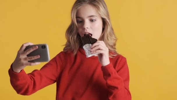 Atraktivní blond dospívající dívka brát selfie na smartphone kousání čokoládové tyčinky přes žluté pozadí. Koncept moderní technologie