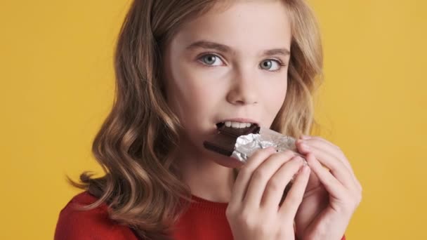 黄色の背景においしいチョコレートバーを食べるかなりブロンドのティーンエイジャーの女の子を閉じます うわ顔 — ストック動画