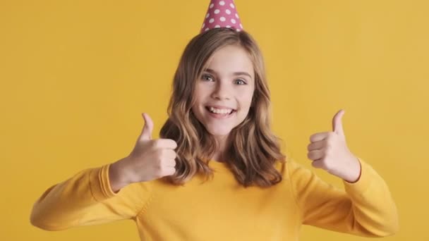 かなり正ブロンドティーン女の子でパーティー帽子見て幸せな維持親指上のカメラの上に黄色の背景 彼女の誕生日パーティーのような陽気な女の子 — ストック動画