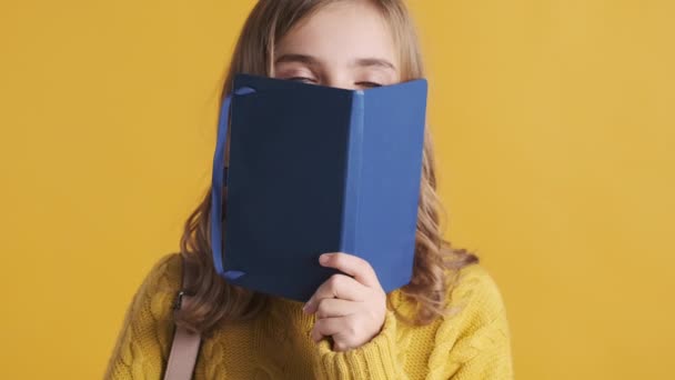漂亮的金发女学生躲在一本书后面偷窥 在黄色背景的相机上嬉闹地摆姿势 有趣的女学生 — 图库视频影像