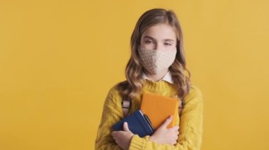 Koruyucu maske takan sarışın öğrenci kız elinde kitaplarla dikilip sarı arka planda kameraya bakıyordu. Güvenlik önce gelir.