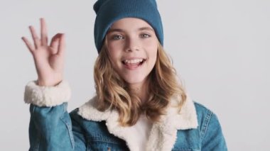 Şapka ve kot ceket giymiş güzel, gülümseyen sarışın kız beyaz arka planda izole edilmiş kamerada iyi bir jest gösteriyor.