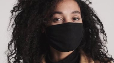 Siyah tıbbi maske takan Afro-Amerikalı kadın beyaz arka plandan kameraya bakıyor. Önce güvenlik kavramı