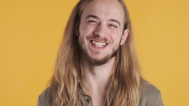 Genç, pozitif sarışın, uzun saçlı, kameraya bakıp gülen, renkli arka planda mutlu görünen bir adam.