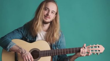 Çekici, uzun saçlı, akustik gitarla oynayan, renkli arka planda kendine güvenen sarışın adam. Stüdyoda gitarist