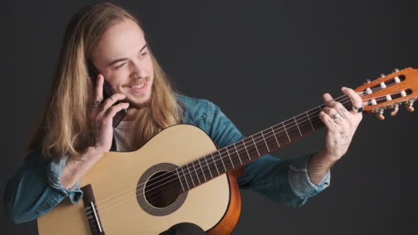 Mladý Dlouhosrstý Muzikant Mluví Smartphonu Zatímco Ladí Kytaru Přes Černé — Stock video