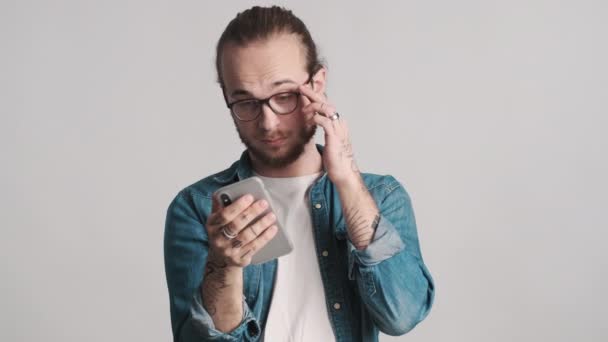 若い集中髭の男は白い背景に隔離された仕事のためのスマートフォンを使用して眼鏡を着用オンライン — ストック動画