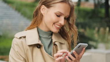 Güzel sarışın kız mutlu görünüyor akıllı telefondan sosyal ağları kontrol ediyor ve dışarıda gülümsüyor. Şehir parkında dinlenen neşeli kız öğrenci.