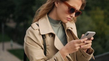 Güneş gözlüğü takan ve trençkot giyen güzel bir öğrenci kız arkadaşlarıyla telefonda konuşurken ciddi görünüyor.