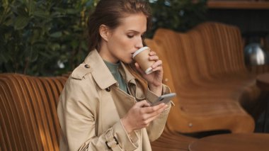 Trençkot giymiş güzel bir öğrenci sokak kafesinde kahve molası sırasında sosyal paylaşım ağı izliyor. Genç bayan kahve içiyor.