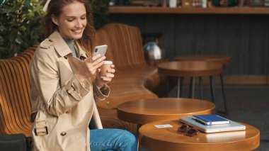 Trençkot giymiş güzel, gülümseyen bir öğrenci kız sosyal paylaşım ağı seyrediyor ve sokak kafesinde ders arası sırasında kahve içiyor.