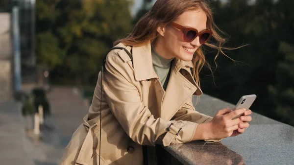 선글라스와 트렌치 코트를 행복해 보이는 아름다운 여학생 공원에서 스마트폰을 사용하여 — 스톡 사진