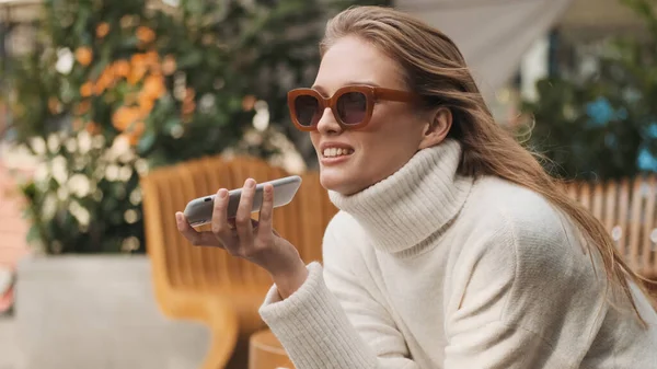 穿着舒适的白色毛衣 戴着太阳镜的漂亮女孩在街上的咖啡店里用智能手机向最好的朋友发送语音讯息 现代技术概念 — 图库照片